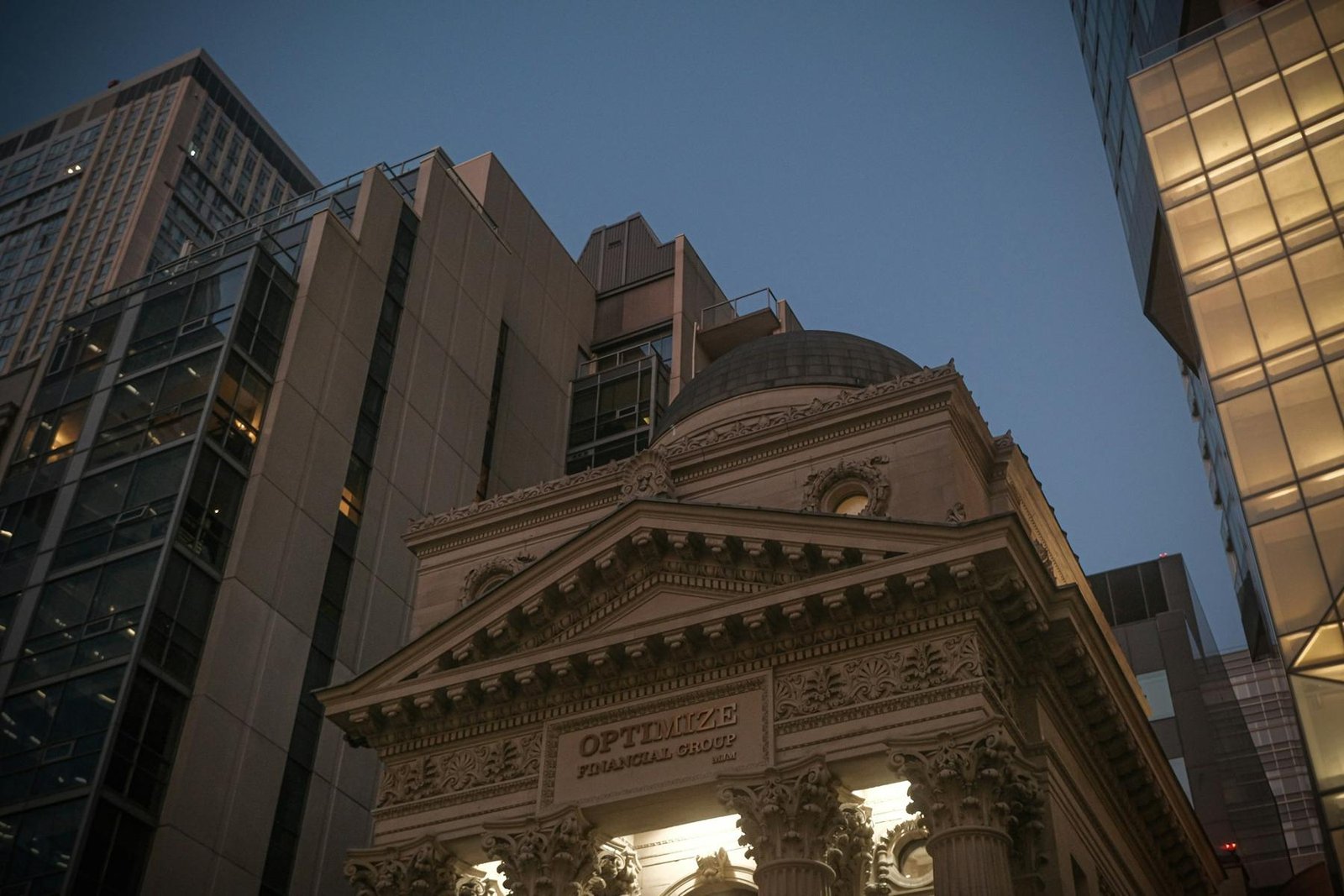 A classical stone bank facade framed by modern office towers at dusk